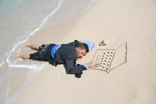 Young man businessman castaway sleeping at the keyboard of his sand computer on empty beach
