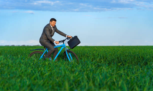 businessman rides a bicycle through a green grass field, dressed in a business suit, he has a briefcase and documents, beautiful nature in spring, business concept stock photo