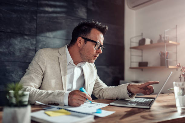 Businessman using laptop and highlighting text in his office Businessman wearing linen suit and eyeglasses sitting at his desk using laptop and highlighting text with blue marker in his office project manager stock pictures, royalty-free photos & images