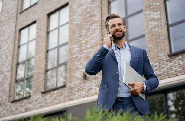 businessman wearing stylish suit talking on mobile phone, holding laptop standing on the street. successful business - makelaar stockfoto's en -beelden