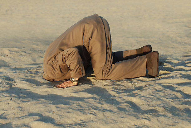 Businessman with head stuck in sand at the beach stock photo