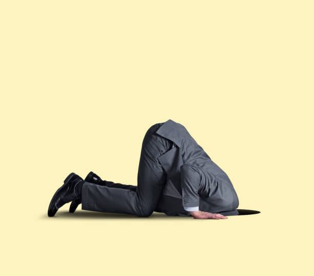 A businessman kneels on all fours as he places his head into a hole in the ground.  Isolated on a yellow background
