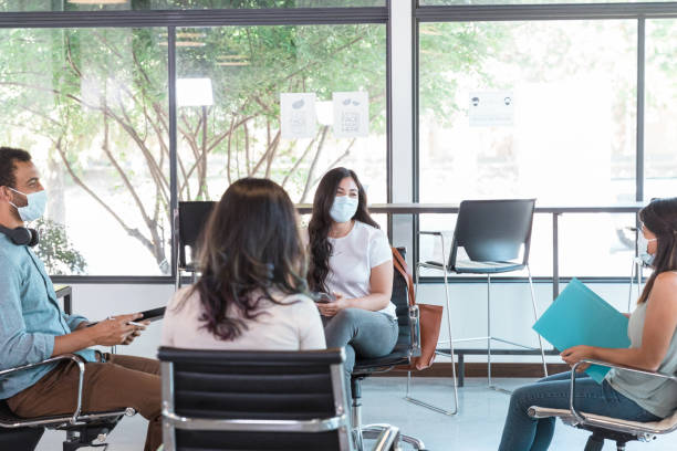 Businesspeople brainstorm ideas A group of business colleagues sit in a circle while participating in a staff meeting. They are wearing protective face masks while practicing social distancing. group therapy stock pictures, royalty-free photos & images