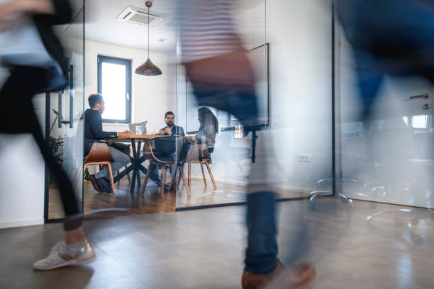Businesspeople in Conference Room and Colleagues Walking By Blurred motion of colleagues walking briskly down office hallway as colleagues sit in conference room discussing ideas. blurred motion stock pictures, royalty-free photos & images