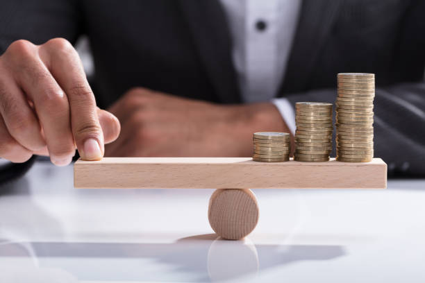 Businessperson Balancing Stacked Coins On Wooden Seesaw Close-up Of A Businessperson's Hand Balancing Stacked Coins On Wooden Seesaw With Finger Over Desk Price and Budget stock pictures, royalty-free photos & images