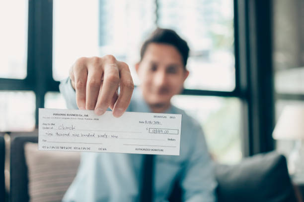 A Businessperson's hand giving cheque to customer and dollar bill, coin, laptop and graph chart on the desk at office. Payment by check, paycheck, payroll concept.  cheque stock pictures, royalty-free photos & images