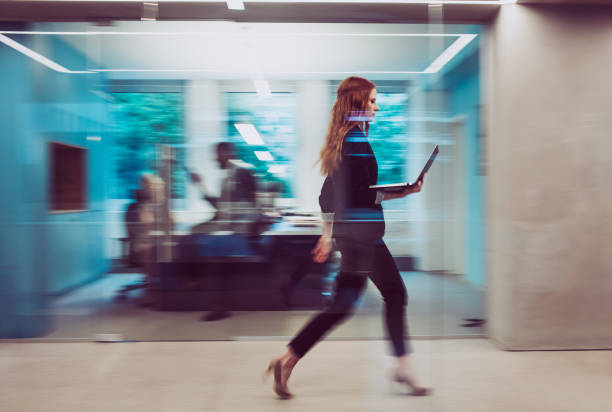 femme d'affaires retenant un ordinateur portatif, marchant en bas du couloir - mobilité photos et images de collection