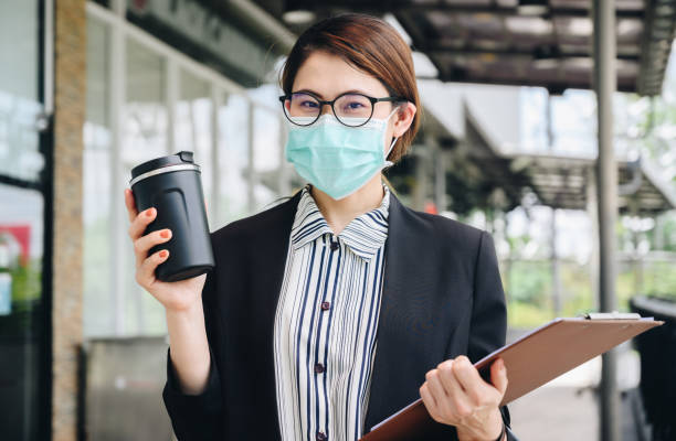Businesswoman holding a tumbler cup of coffee and wearing mask for protect herself in covid-19 pandemic while walking on city street to her office. stock photo