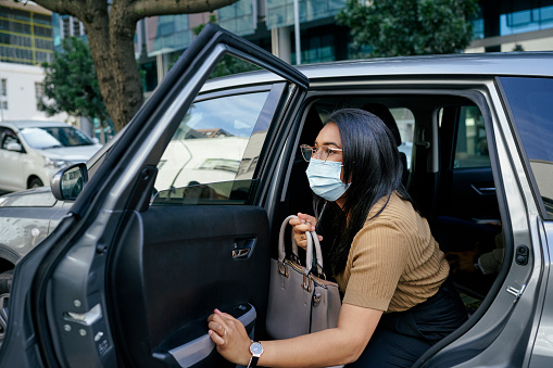 Young businesswoman wearing a protective face mask getting out of the back seat of a taxi on a city street