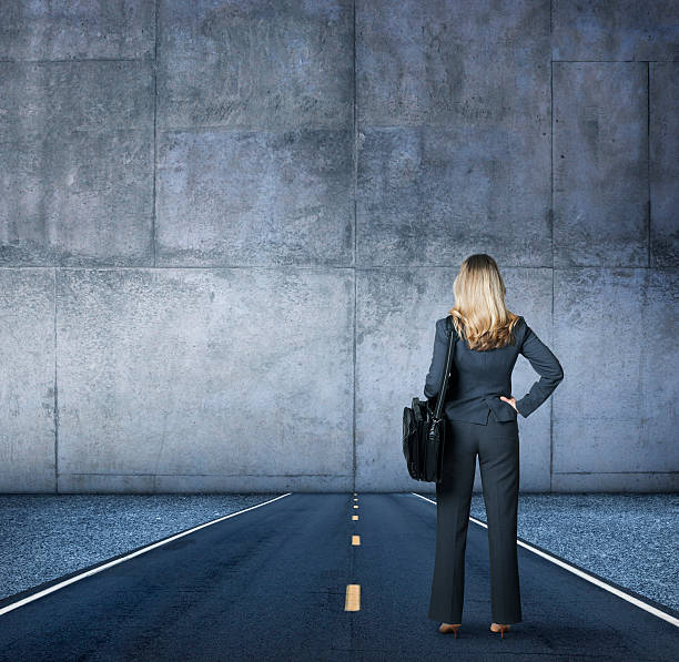 Businesswoman Looking At The End Of The Road A businesswoman, with her back to the camera while holding a shoulder bag, stands in the middle of a road that runs directly into a large concrete wall. dead end road stock pictures, royalty-free photos & images