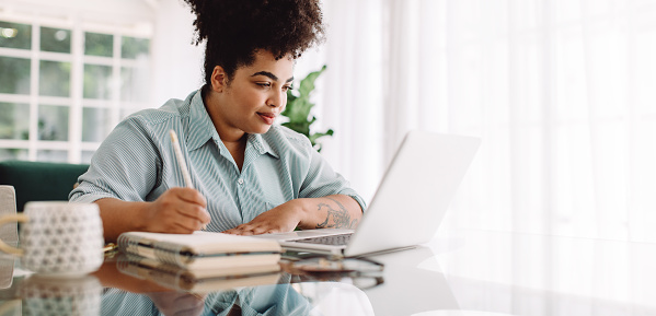 Business woman working from home writing notes while looking at laptop. Confident woman sitting at desk using laptop and taking notes.