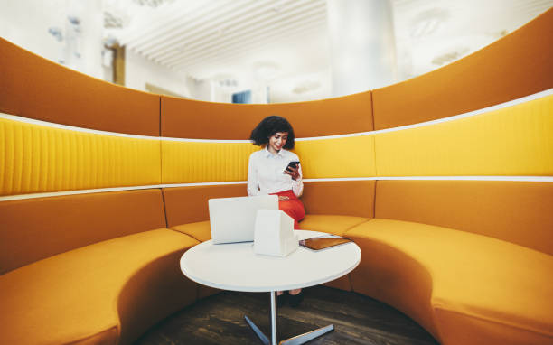 Businesswoman on a yellow sofa A wide-angle shot of a young elegant African-American businesswoman using her smartphone and gadgets while sitting on a round yellow sofa in office coworking area; woman entrepreneur with a cellphone soundproof stock pictures, royalty-free photos & images