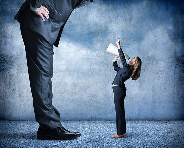 Businesswoman Shouts Through Megaphone Towards Much Larger Businessman A businesswoman shouts through a megaphone and points up towards a much larger businessman who is standing over her. assertiveness stock pictures, royalty-free photos & images