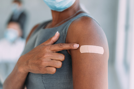 Cropped shot of an attractive african-american businesswoman pointing at her arm with a bandage after receiving COVID-19 vaccine in the office with her colleagues in background. Young businesswoman showing her shoulder after getting coronavirus vaccine.