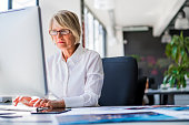 A photo of mature businesswoman using computer at desk. Concentrated female executive is working on desktop PC. Confident female professional is in formals. She is in brightly lit office.