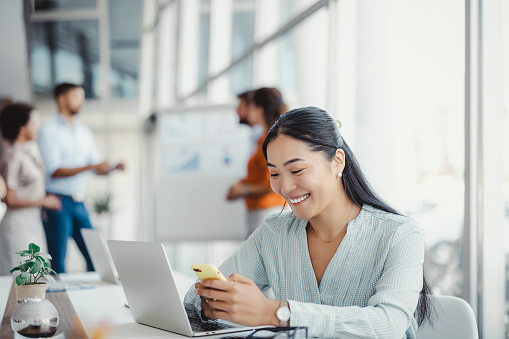 Young smiling Asian business woman using smartphone near computer in office with colleagues in the background, copy space