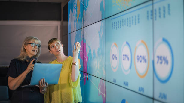 Businesswomen Discussing Ideas Against an Information Wall An experienced woman mentors a female colleague, the mature woman is holding a laptop as they debate data from an interactive display; they are both wearing smart casual clothing. business strategy photos stock pictures, royalty-free photos & images