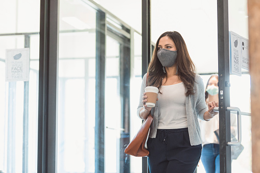 A mid adult businesswoman enters her office during the COVID-19 pandemic. A colleague is walking behind her, practicing the proper social distancing protocol. The businesswomen are wearing protective face masks.