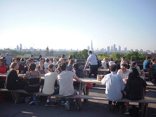 Busy Rooftop Bar in London on Hot Summer's Day Peckham, London, UK – June 12, 2015: Young adults eating and drinking on a rooftop bar during a hot London summer's day. The bar is on top of a car park in Peckham, SE London, serves food, beers, wine and cocktails, and is always incredibly popular during the summer in London. bar drink establishment stock pictures, royalty-free photos & images