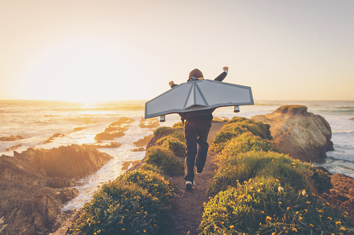 A young boy dressed in business suit and tie wears a homemade jetpack and flying goggles raises his arms in the afternoon sun while running to take off into the air on an outcropping above the surf in California. This young entrepreneur is ready to take his new business to new heights.