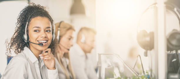 Call center worker accompanied by her team. Call center worker accompanied by her team. Smiling customer support operator at work. Young employee working with a headset. service stock pictures, royalty-free photos & images