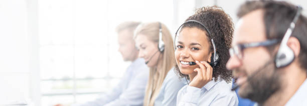 Call center worker accompanied by her team. Call center worker accompanied by her team. Smiling customer support operator at work. Young employee working with a headset. customer service representative stock pictures, royalty-free photos & images
