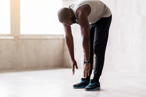 Calm african muscular guy doing warming-up exercises Concentrated bearded sportsman is standing on straight legs and making forward bend. Focus on smart watch and sport shoes bending stock pictures, royalty-free photos & images