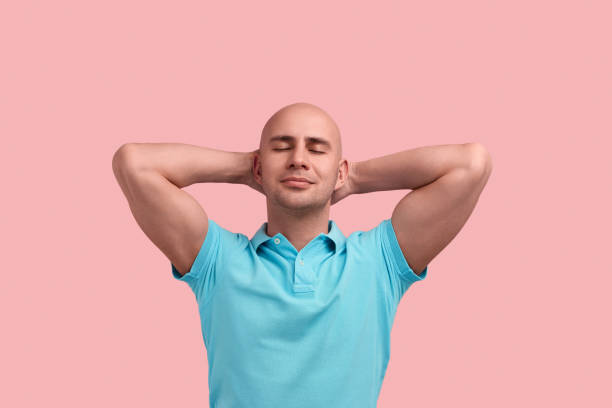 毛を持つ穏やかなハゲ同性愛者の男はリラックスしている、目を閉じて、頭の下に手を保つ、地面に横たわっている、太陽を楽しむ、ゲイフレンドリー、青いポロシャツを着て、ピンクの背� - 快感 ストックフォトと画像