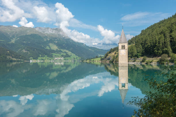 Calm water of the lake Resia with drowned bell tower and magical reflections of the sky and the mountains in it stock photo