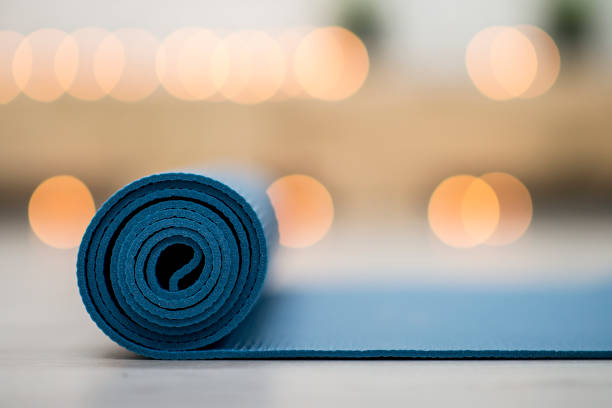 Sebuah tikar yoga biru sedang diluncurkan ke lantai putih di dalam studio yoga. Ini adalah malam dan ada lampu lilin buram di belakang tikar.