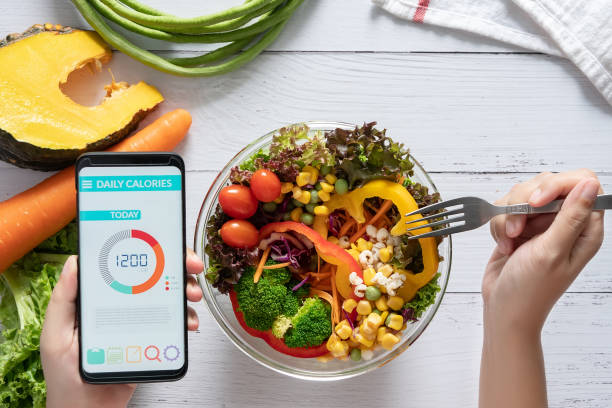 Calories counting , diet , food control and weight loss concept. Calorie counter application on smartphone screen at dining table with salad, fruit juice, bread and fresh vegetable. healthy eating stock photo