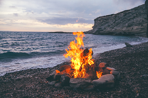 Summer Camp fire burning in stones on the pebble beach and against sea coast line, rocks and sunset sky.