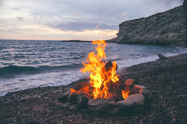Campfire on the beach Summer Camp fire burning in stones on the pebble beach and against sea coast line, rocks and sunset sky. bonfire stock pictures, royalty-free photos & images