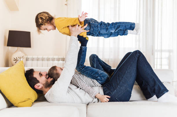 I can fly with your support! Close-up of a beautiful children playing with their father in domestic room. Little girl is flying up in the air. fathers day stock pictures, royalty-free photos & images