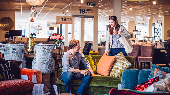 Young women out shopping with her husband in a furniture shop. They are enjoying looking at the sofas and cushions. The man is sitting on one of the sofas as the woman smiles standing looking at him.