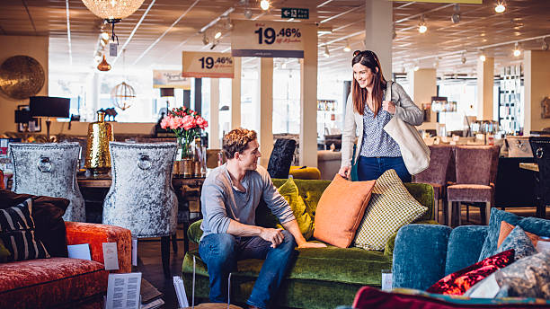 Can we buy this sofa? Young women out shopping with her husband in a furniture shop. They are enjoying looking at the sofas and cushions. The man is sitting on one of the sofas as the woman smiles standing looking at him. furniture stock pictures, royalty-free photos & images