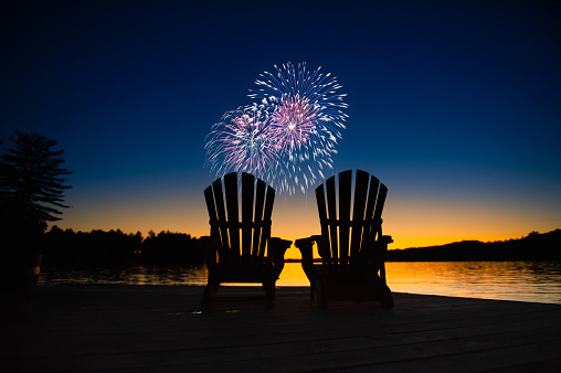 Canada day fireworks on a lake in Muskoka, Ontario Canada. On the wooden dock two Adirondack chairs are facing the sunset orange hues