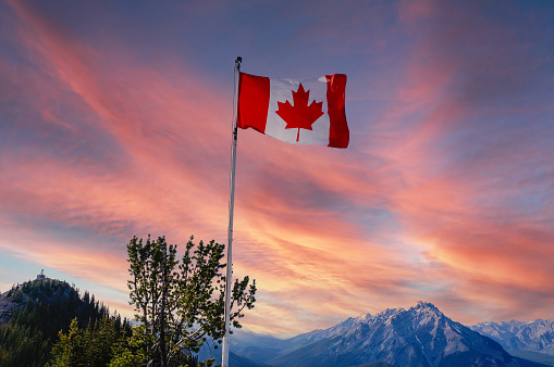 Canada flag flying atop Sulphur Mountain with sunset view of Banff National Park with Canadian Rockies in the background.