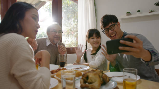 Candid of happy asian family having fun on dining table at home holding mobile video call online and selfie or take photo shoot group with smile and laugh together. Candid of happy asian family having fun on dining table at home holding mobile video call online and selfie or take photo shoot group with smile and laugh together. asian family eating together stock pictures, royalty-free photos & images