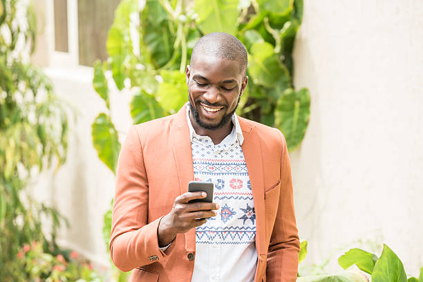 Candid portrait of Nigerian man using cell phone, smiling Young African man in orange jacket using smartphone. Young happy Ethnic man texting on phone. Cheerful casual male reading text message. nigeria stock pictures, royalty-free photos & images