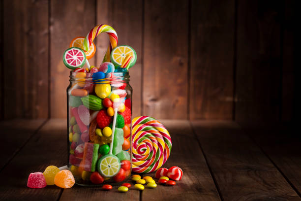 Candy jar on rustic wooden table Front view of an open candy jar filled with multi colored candies, lollipops and jelly beans standing on rustic wooden table. Some candies are spilled out of the jar directly on the background. DSRL low key studio photo taken with Canon EOS 5D Mk II and Canon EF 100mm f/2.8L Macro IS USM candy jar stock pictures, royalty-free photos & images