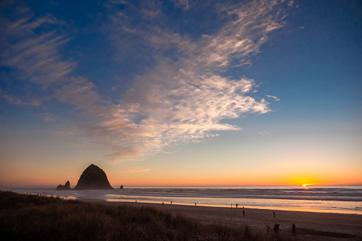 Oregon's Iconic Cannon Beach at sunset. Haystack Rock rises out of the sandy surf as cotton candy clouds float weightlessly in the orange pink and blue sky and the sun sets just over the horizon of the Pacific Ocean.