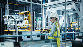 istock Car Factory: Female Automotive Engineer Wearing Hard Hat, Standing, Using Laptop. Monitoring, Control, Equipment Production. Automated Robot Arm Assembly Line Manufacturing Electric Vehicles. 1352825077