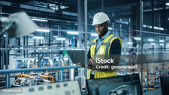 istock Car Factory: Professional Male Automotive Engineer Wearing Hard Hat, Walking, Using Laptop. Monitoring, Control, Equipment Production. Automated Robot Arm Assembly Line Manufacturing Electric Vehicles 1352825038