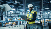 istock Car Factory: Professional Male Automotive Engineer Wearing Hard Hat, Walking, Using Laptop. Monitoring, Control, Equipment Production. Automated Robot Arm Assembly Line Manufacturing Electric Vehicles 1352825038