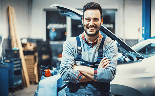 Car mechanic at work. Closeup front view portrait of smiling handsome mechanic standing in front of a car with his arms folded, smiling and looking at camera. mechanic stock pictures, royalty-free photos & images