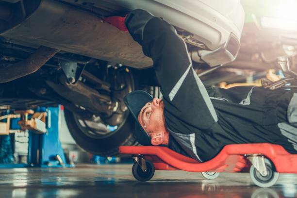 Car Mechanic Under the Car Caucasian Car Mechanic in His 30s Under the Car on the Mechanics Creeper Trying To Fix Modern Vehicle Exhaust System. below stock pictures, royalty-free photos & images