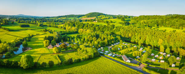 Aerial panorama over caravan trailer park campsite nestled in a picturesque river valley meandering between rolling hills of patchwork pasture and green summer landscape.