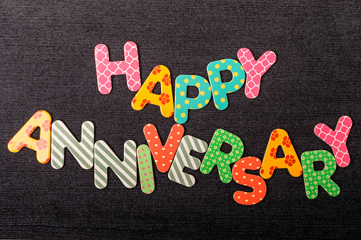 Card with Happy Anniversary words made from mixed vivid colored wooden letters on a textured dark black textile material that can be used as a message