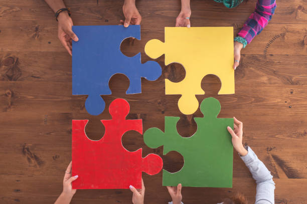 Cardboard puzzle pieces Overhead view of four large puzzle pieces that fit together into a square. There are four students who are each holding a piece. The pieces are blue, yellow, red, green. The background is a wooden table. four people stock pictures, royalty-free photos & images